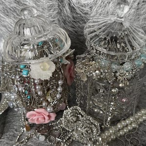 May include: Two clear glass jars filled with jewelry, including necklaces, bracelets, and rings. The jewelry is silver-toned with pearls, turquoise, and floral accents. The jars have decorative lids and sit on a gray surface.