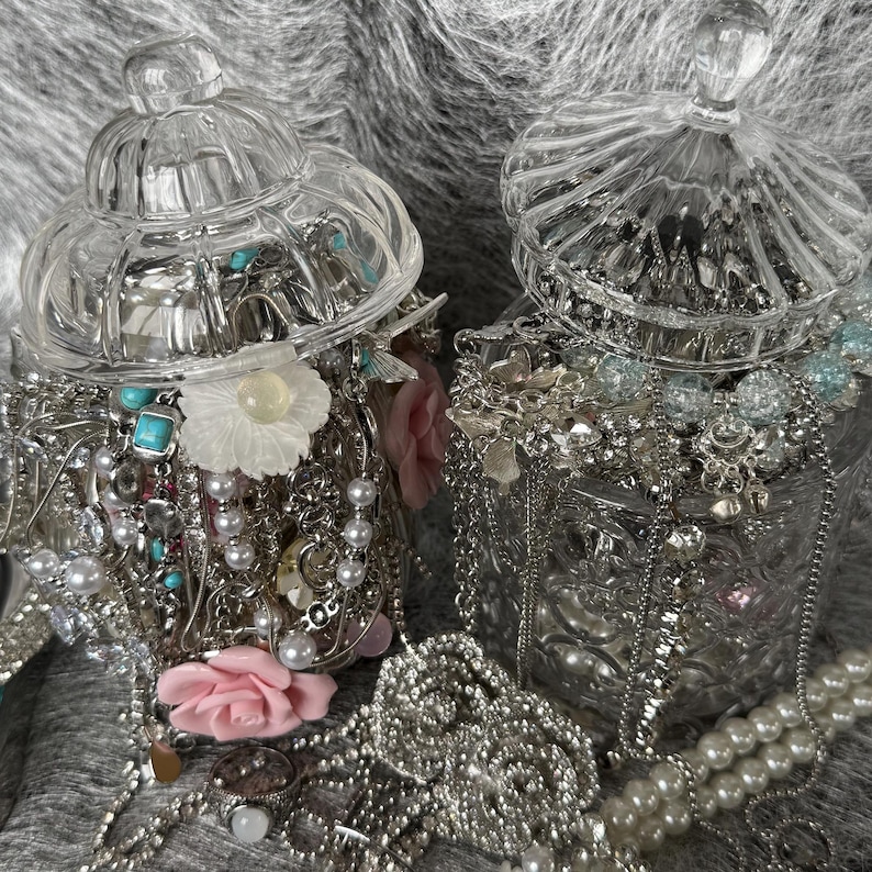 May include: Two clear glass jars filled with jewelry, including necklaces, bracelets, and rings. The jewelry is silver-toned with pearls, turquoise, and floral accents. The jars have decorative lids and sit on a gray surface.