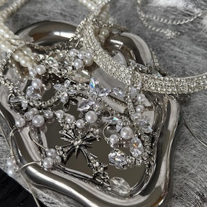 May include: A collection of silver-toned jewelry, including necklaces, bracelets, and brooches, displayed on a silver tray. The jewelry features pearls and clear crystals. The tray sits on a gray, textured surface.