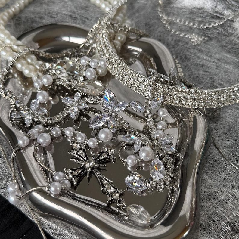May include: A collection of silver-toned jewelry, including necklaces, bracelets, and brooches, displayed on a silver tray. The jewelry features pearls and clear crystals. The tray sits on a gray, textured surface.