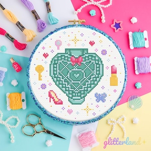 May include: A cross-stitch embroidery hoop featuring a teal perfume bottle design with a pink heart, surrounded by small icons. The background includes colorful embroidery floss, scissors, and the text "glitterland +."