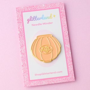 May include: A peach-colored needle minder with a gold outline, shaped like a vintage toy. The minder features the text "Polly Pocket" in a circular design. The background is a pink glitter card with the text "glitterland + Needle Minder".