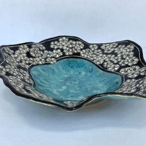 May include: A shallow ceramic bowl with a wavy, irregular rim. The center features a textured, mottled turquoise glaze, while the wide, scalloped border is finished in black with a repeating white hand-drawn floral pattern of small five-petaled flowers.