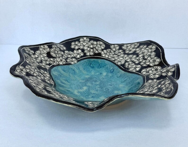 May include: A shallow ceramic bowl with a wavy, irregular rim. The center features a textured, mottled turquoise glaze, while the wide, scalloped border is finished in black with a repeating white hand-drawn floral pattern of small five-petaled flowers.