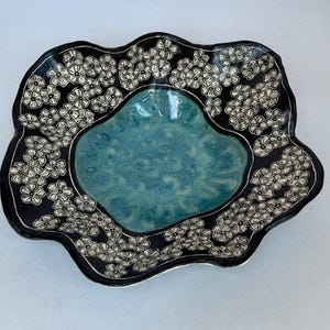 May include: A ceramic bowl with a wavy, irregular rim. The center features a glossy, mottled turquoise glaze, while the wide, dark black border is decorated with a repeating pattern of small, white, hand-drawn five-petaled flowers.