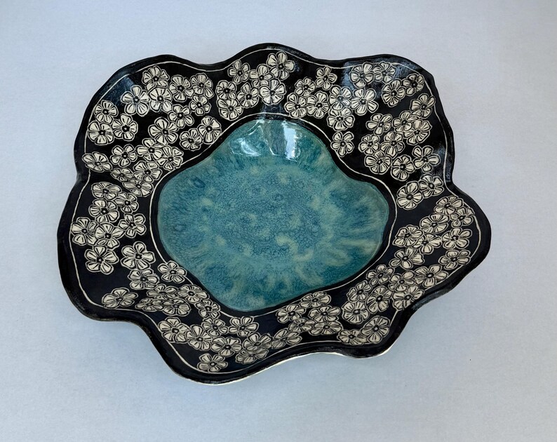 May include: A ceramic bowl with a wavy, irregular rim. The center features a glossy, mottled turquoise glaze, while the wide, dark black border is decorated with a repeating pattern of small, white, hand-drawn five-petaled flowers.