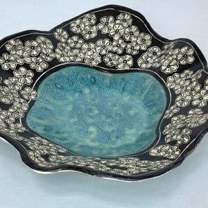 May include: A ceramic bowl with a wavy, irregular rim. The center features a textured, mottled turquoise glaze, while the wide, dark black border is decorated with a repeating pattern of white, hand-drawn five-petaled flowers and leaves.