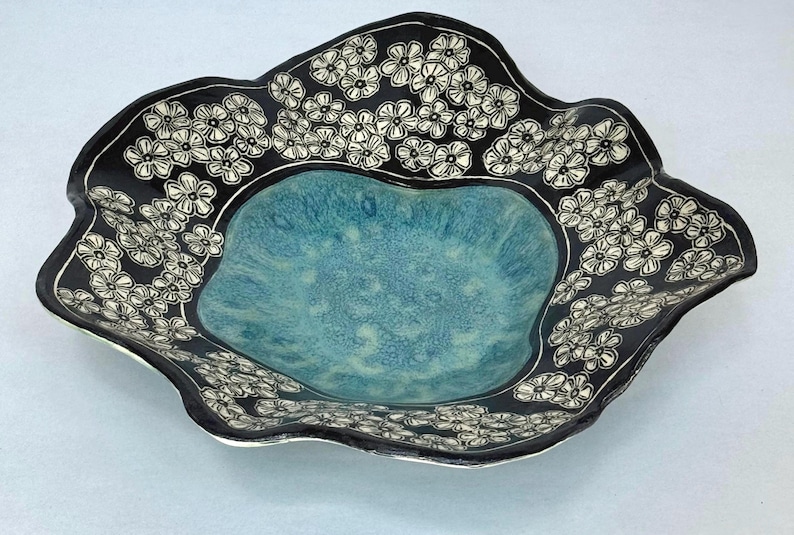 May include: A ceramic bowl with a wavy, irregular rim. The center features a textured, mottled turquoise glaze, while the wide, dark black border is decorated with a repeating pattern of white, hand-drawn five-petaled flowers and leaves.