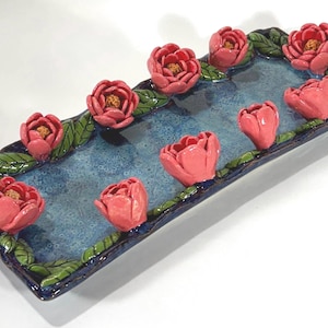 May include: A rectangular ceramic serving dish with a blue interior and a dark blue border. The border is decorated with green leaves and pink flower sculptures. The dish is approximately 12 inches long.