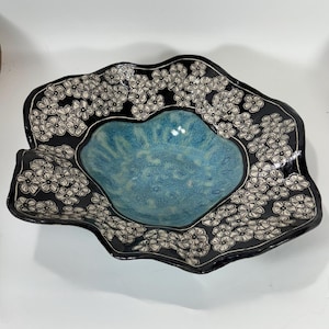 May include: A decorative ceramic bowl with a unique, wavy shape. The bowl features a black rim adorned with white floral patterns. The interior is a vibrant blue, creating a striking contrast. This handcrafted piece is ideal for serving or display.