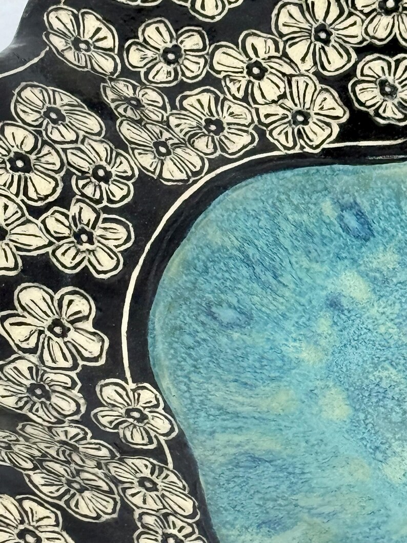 May include: A ceramic bowl featuring a central basin with a textured, mottled turquoise and light blue glaze. The wide, dark black rim is decorated with a repeating pattern of carved, cream-colored five-petaled flowers with dark centers.
