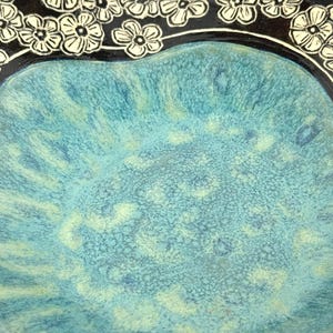 May include: A ceramic bowl featuring a vibrant turquoise and blue mottled glaze in the center, surrounded by a dark black border decorated with a repeating pattern of white five-petaled flowers with detailed line work throughout the petals.