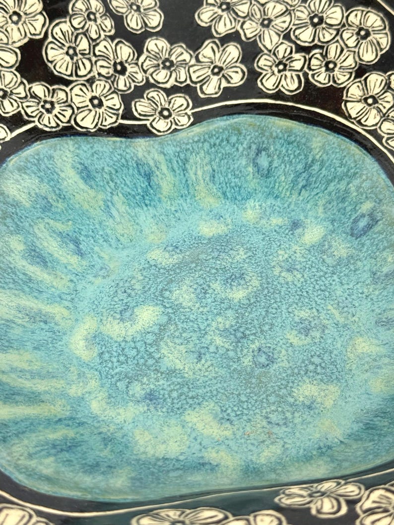May include: A ceramic bowl featuring a vibrant turquoise and blue mottled glaze in the center, surrounded by a dark black border decorated with a repeating pattern of white five-petaled flowers with detailed line work throughout the petals.