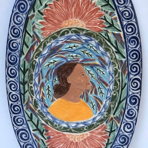 May include: Oval ceramic plate with a detailed floral and figure design. The plate features a blue border with spiral patterns, surrounding a central image of a woman in a yellow top, with a tear. The design includes orange flowers and green leaves.