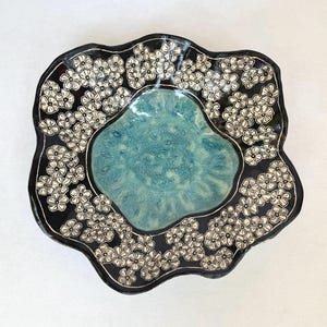 May include: A ceramic bowl with an irregular, wavy rim. The center features a smooth, mottled turquoise glaze, while the wide border is finished in a glossy black glaze decorated with a repeating pattern of small, white, five-petaled flowers.