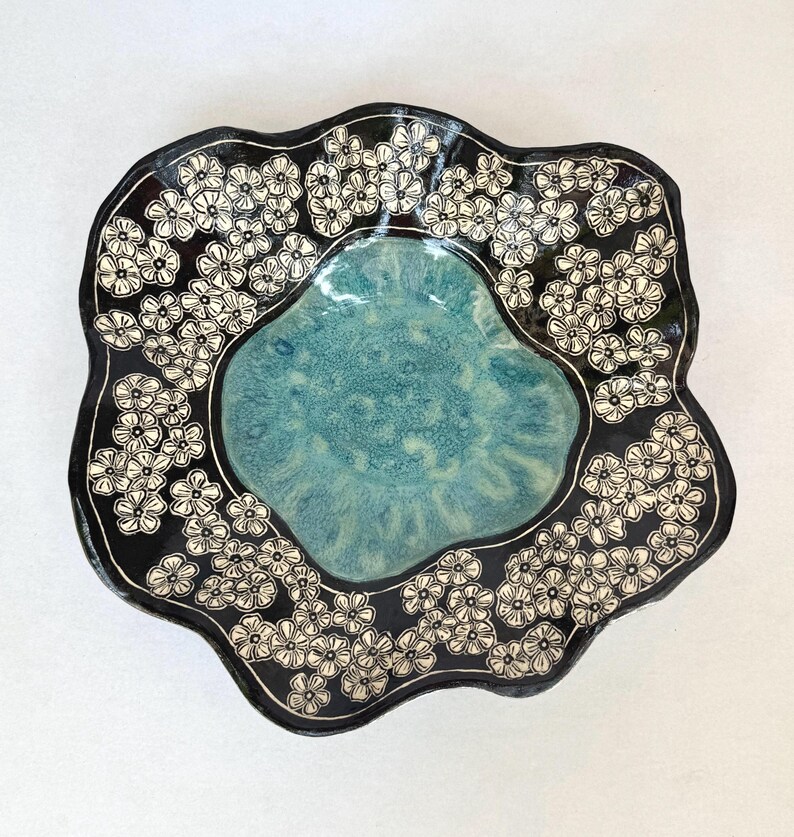 May include: A ceramic bowl with an irregular, wavy rim. The center features a smooth, mottled turquoise glaze, while the wide border is finished in a glossy black glaze decorated with a repeating pattern of small, white, five-petaled flowers.