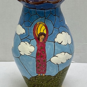 May include: A ceramic vase with a ruffled purple rim and a blue sky background featuring white clouds. The body depicts a figure in a magenta dress with green leaf patterns standing on a green hill, holding a yellow sun overhead in a mosaic style.