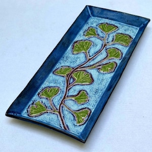 May include: A rectangular ceramic tray featuring a deep blue border and a light blue center with a textured, woven-like pattern. A central branch design displays multiple green ginkgo leaves with dark brown outlines, creating a botanical motif.