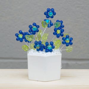 Puede incluir: Un arreglo floral decorativo con flores de cuentas azules con detalles en amarillo y verde. Las flores están dispuestas en una maceta de cerámica blanca cuadrada, creando una exhibición encantadora y colorida. El arreglo está sobre un fondo neutro.