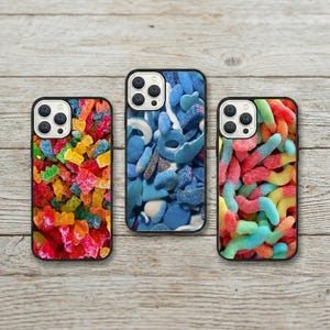 Op de afbeelding: Drie telefoonhoesjes met zwarte randen en kleurrijke snoepontwerpen. Eén hoesje heeft een mix van gummyberen, een ander blauwe ringvormige snoepjes en de derde toont gummy wormen in verschillende kleuren.
