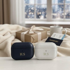 May include: Two personalised AirPod cases, one navy blue and one white, each with the initials "H.S." in gold lettering. A wrapped gift, passport, and mug are in the background, suggesting a travel theme.