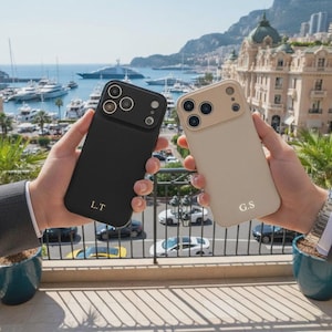 Puede incluir: Dos fundas de teléfono, una negra y otra beige, se sostienen contra un fondo escénico. La funda negra tiene las iniciales "L.T." y la funda beige "G.S.". Ambas fundas tienen una protuberancia para la cámara.