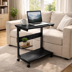May include: Black rolling laptop desk with a small shelf and a lower storage area. A laptop and a mug sit on the top surface. A small plant and a basket are on the shelf. The desk is next to a beige sofa.
