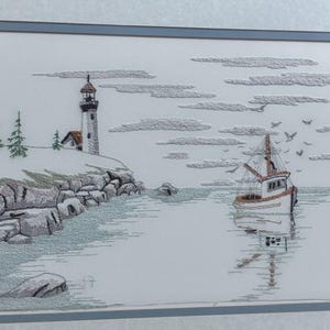 May include: A framed embroidery art piece depicting a coastal scene with a lighthouse on a rocky shore, green pine trees, and a small fishing boat on the water. The sky features textured gray clouds and a flock of birds, all rendered in thread.