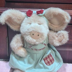 May include: A plush pig toy with large, floppy ears and a red bow. The pig is wearing a light blue dress with a red and brown plaid pocket. Text on the dress reads "This little piggy went to the market..."