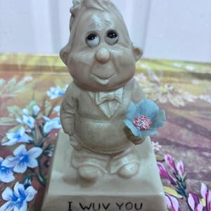 May include: A beige figurine of a cartoonish man holding a blue flower with a pink center. The figurine is standing on a rectangular base with the words "I WUV YOU" in black lettering.