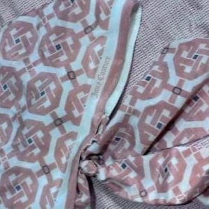 May include: A patterned scarf in shades of pink, white, and navy blue, with a repeating geometric design. The scarf has a light blue border with the text "Juicy Couture". The scarf is made of a soft, flowing fabric.