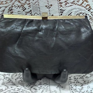 May include: A black leather clutch with a gold-toned clasp. The rectangular bag has a textured surface and a simple, elegant design. The clutch is displayed on a black stand.