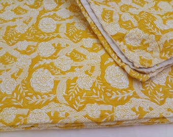 May include: A folded yellow and white floral patterned quilt. The quilt features a repeating design of large white flowers and winding vines on a yellow background. The edges are neatly finished.