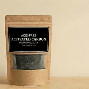 Op de afbeelding: Een bruine papieren zak met een doorzichtig venster, met actieve kool. De zak heeft een zwart etiket met witte tekst: "ACID-FREE ACTIVATED CARBON PREMIUM QUALITY FOR 3D PRINTER". De achtergrond is lichtbeige.