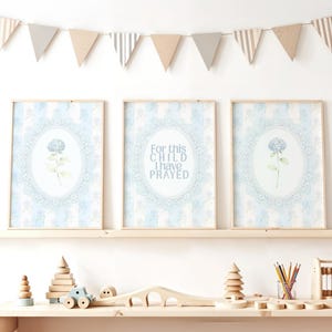 Puede incluir: Tres impresiones enmarcadas con un diseño floral azul claro y el texto "For this child I have prayed." Las impresiones se exhiben en un estante de madera con juguetes de madera y una pancarta de banderas triangulares.