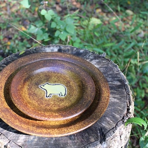 May include: Two brown ceramic plates stacked on a tree stump. The top plate has a light green bear sticker in the center. The plates have a speckled, rustic appearance, with a slightly purple hue in the center.