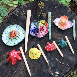 May include: A collection of ceramic mushroom-shaped sculptures and pointed wooden sticks. The ceramic pieces feature various colors including green, yellow, red, orange, and a spiderweb pattern. The wooden sticks are light-colored and tapered.