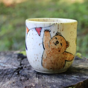 May include: A cream-colored ceramic mug with a speckled finish featuring a hand-drawn illustration of a brown bear reaching up toward a string of colorful triangular bunting flags. The bear has a simple face and is rendered in warm brown tones.