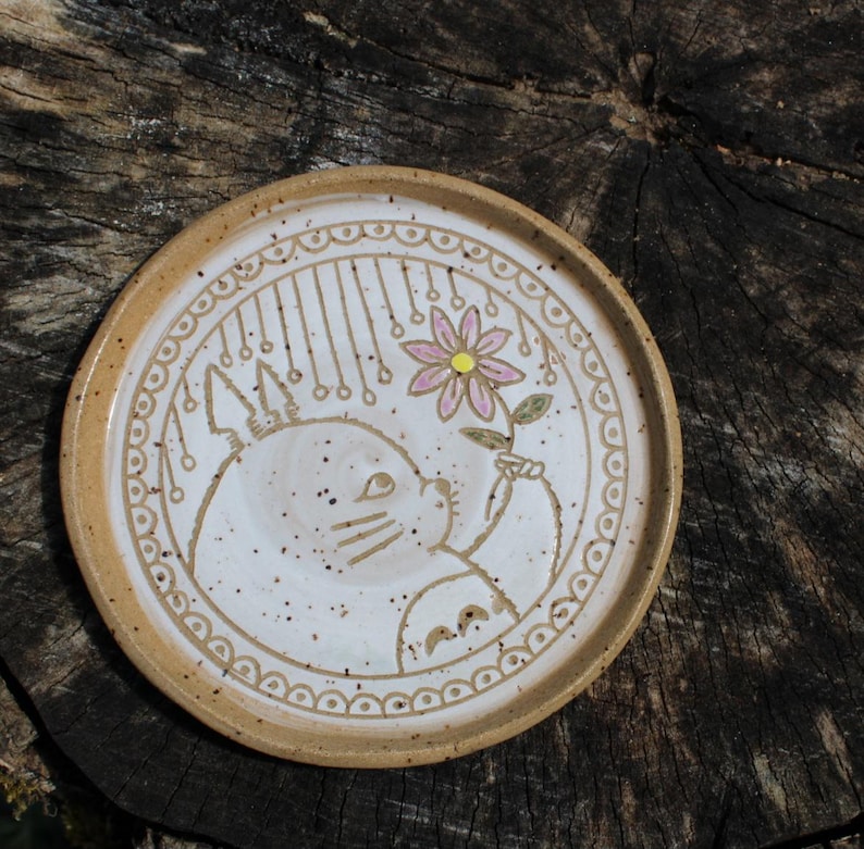 May include: A round, ceramic plate with a beige rim and a white center. The center features a hand-drawn illustration of a cat holding a flower. The cat is surrounded by a decorative border and hanging lines.