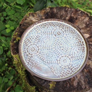 May include: A round ceramic plate featuring a white lace doily impression on a light blue and brown glazed surface. The plate has a smooth, glossy finish with a dark brown rim, showcasing a detailed, intricate pattern pressed into the clay.