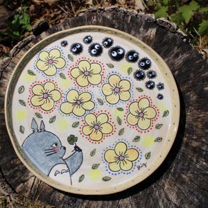 May include: A round, hand-painted ceramic plate featuring a whimsical design. The plate showcases yellow flowers, green leaves, and black soot sprites. A gray cartoon character holds a four-leaf clover. The plate has a cream-colored background and a brown rim.