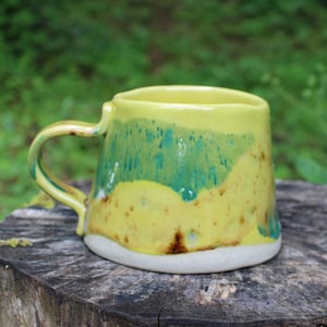 May include: A ceramic mug with a glossy finish featuring a bright yellow upper section that transitions into a vibrant teal drip pattern. The base is a matte, unglazed off-white color, and the mug includes a curved handle on the left side.