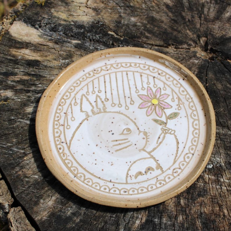 May include: A ceramic plate with a hand-drawn illustration of a cartoon character holding a flower. The plate has a speckled white center, a tan rim, and a decorative border. The flower is pink and yellow.