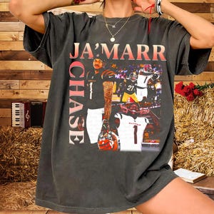 Puede incluir: Camiseta gris oscuro con un gráfico de un jugador de fútbol americano, el nombre "JA'MARR CHASE" en rojo y blanco. La camiseta incluye imágenes del jugador en acción, con el número 1 en un lugar destacado.