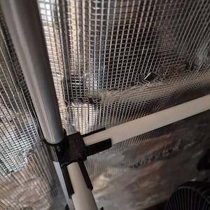 May include: Interior of a grow tent with a reflective silver lining and a framework of white and grey poles. Black connectors join the poles, creating a structured environment. A black fan is visible in the lower right corner.