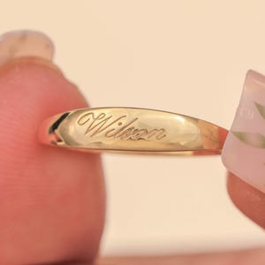 Pode incluir: Um anel de ouro com o nome "Wilson" gravado. O anel é segurado entre dois dedos. O anel tem uma superfície lisa e polida com o nome gravado em uma fonte cursiva.