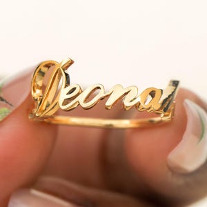 Puede incluir: Un anillo dorado con el nombre "Deonai" en fuente cursiva. El anillo se sostiene sobre un fondo claro. Es una pieza de joyería.