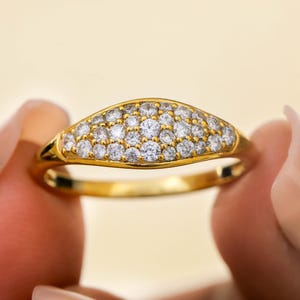 May include: A gold ring featuring a curved, oval-shaped top, set with many small, clear gemstones. The ring is held between two fingers against a soft, yellow backdrop. The ring's band is a simple, polished gold.