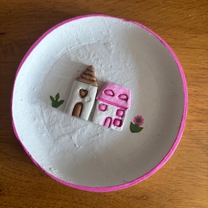 May include: A handcrafted, circular, white dish with a pink border. Two miniature houses are in the middle, one with a brown roof and the other with a pink roof. Green leaves and a pink flower are painted on the dish.