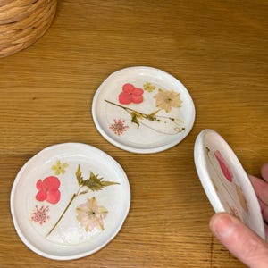 May include: Three white ceramic coasters with dried flowers set in resin. The coasters display a variety of colourful flowers, including pink, yellow, and white blooms. The coasters are round and have a slightly raised edge.